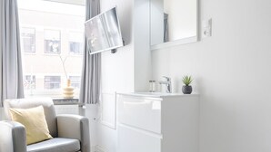 Un baño moderno con tocador blanco, televisor montado y ventana con cortinas.