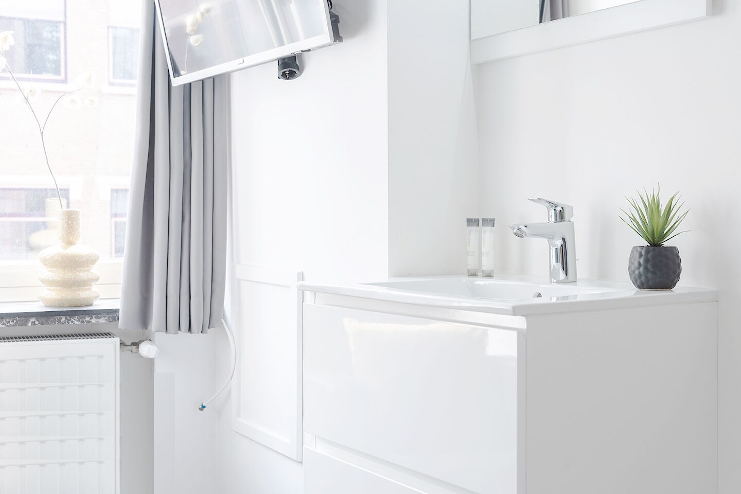 A modern bathroom with a white sink, a mirror, a potted plant, and a window with curtains.