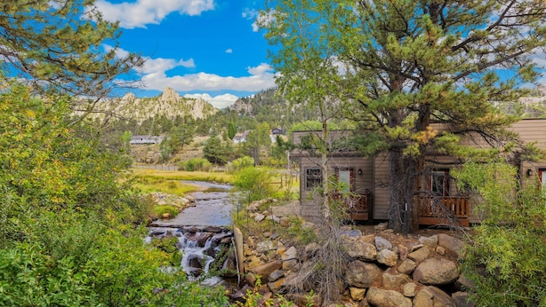 A rustic cabin with a small waterfall, surrounded by lush greenery and a mountainous backdrop.