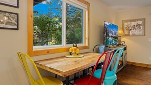 A dining area with a wooden table, colorful chairs, a window with a view of trees, and a television on a stand.
