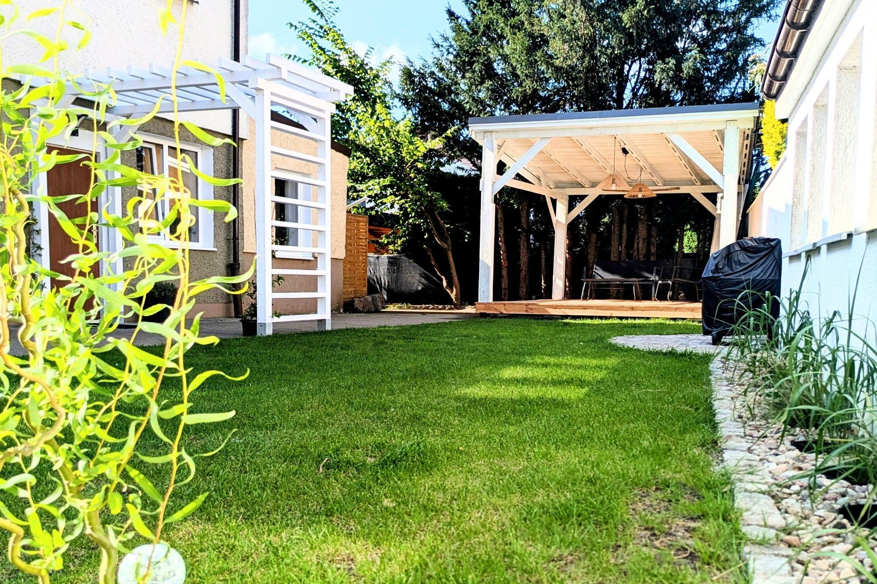 Un patio trasero con un porche techado, una pérgola blanca y un césped cuidado.