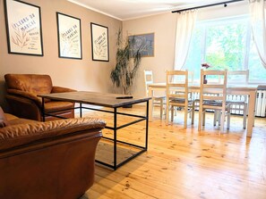 A living room with a brown leather sofa, a wooden coffee table, a dining table with chairs, and framed artwork on the wall.