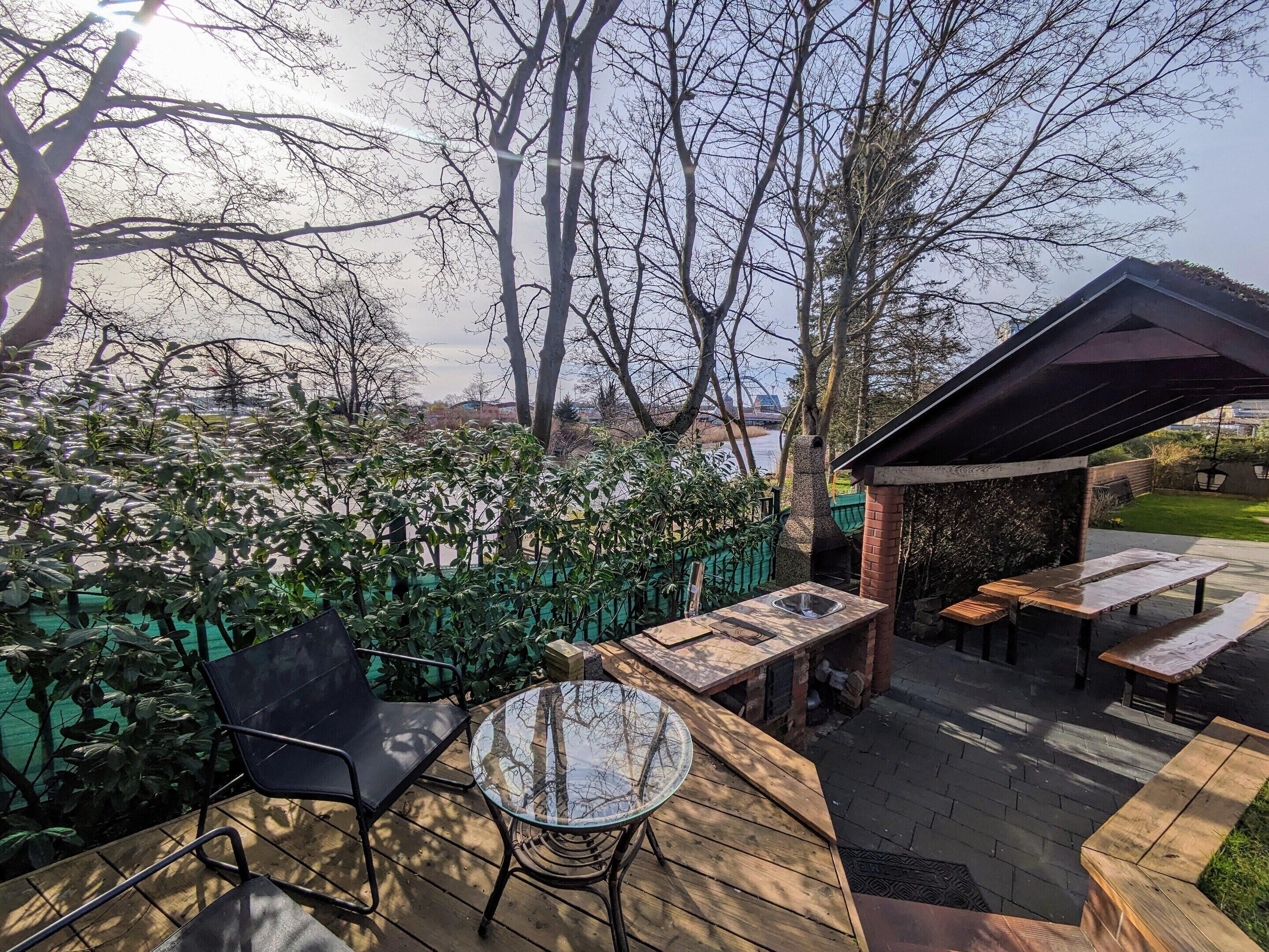 An outdoor patio area with a round glass table and chairs, a brick barbecue, and a covered seating area with benches.