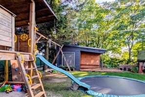 A wooden playhouse with a slide, a trampoline, and a small shed in a backyard. - Large Holiday Apartment With Sauna (Kołobrzeg)