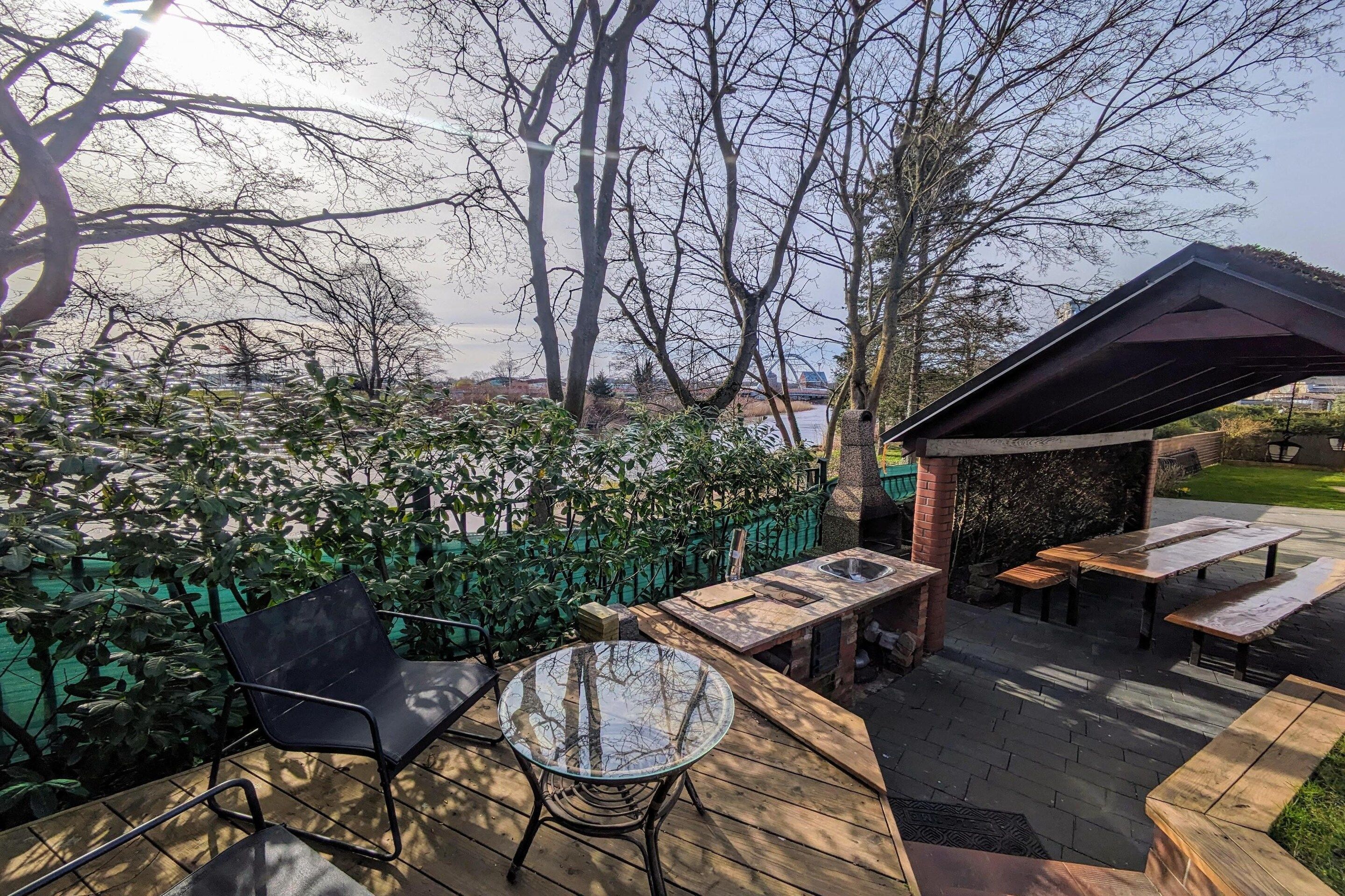 Une terrasse extérieure avec une table et des chaises en verre, entourée d’une plateforme en bois et d’une clôture verte.