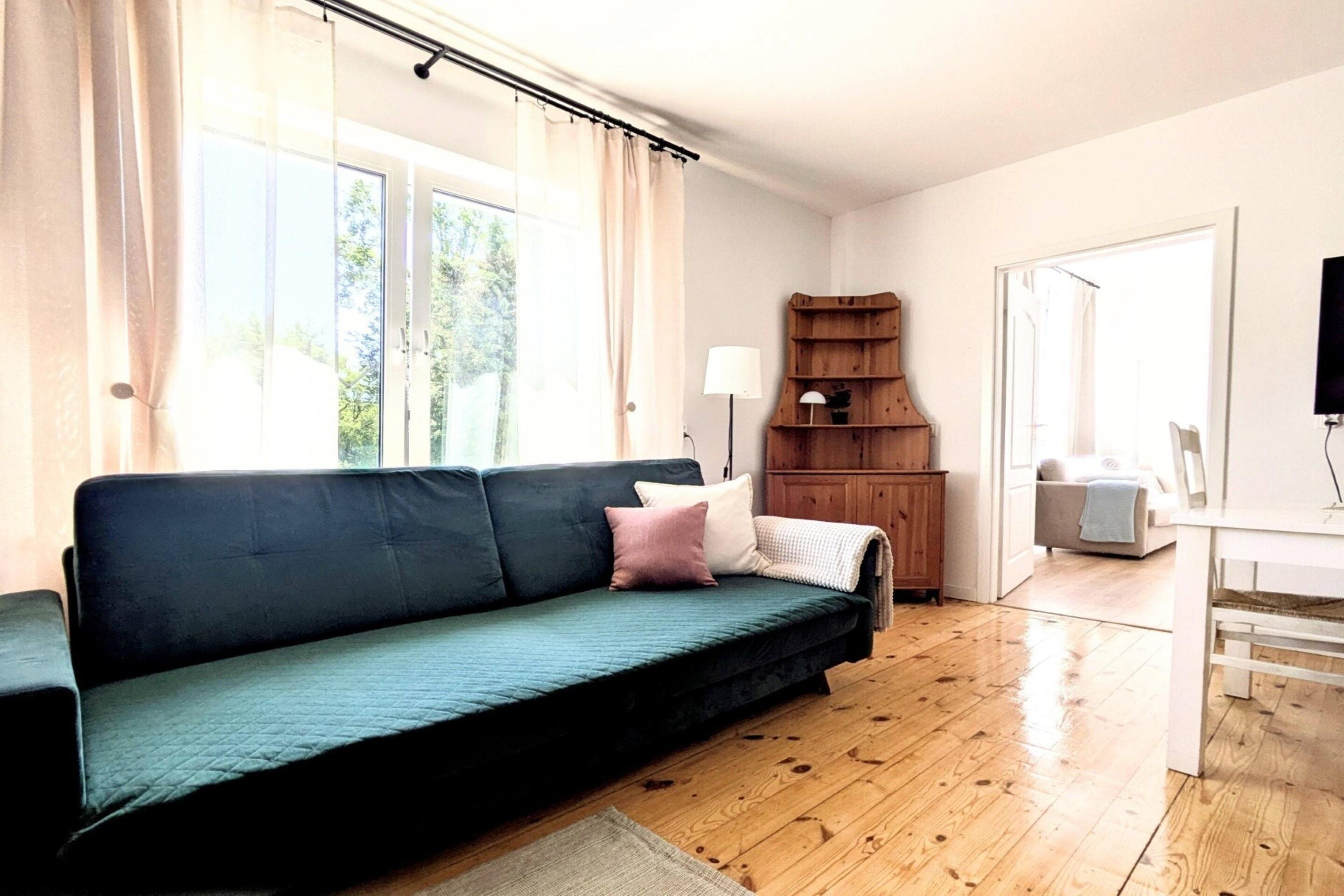 Een kamer met een donkergroene bank, een houten boekenkast, een klein tafeltje met een lamp en een televisie op een standaard.