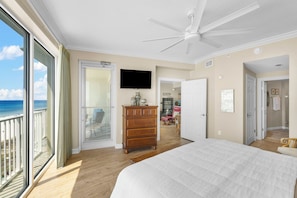 A bedroom with a large bed, a ceiling fan, a dresser, a TV, and a view of the ocean.