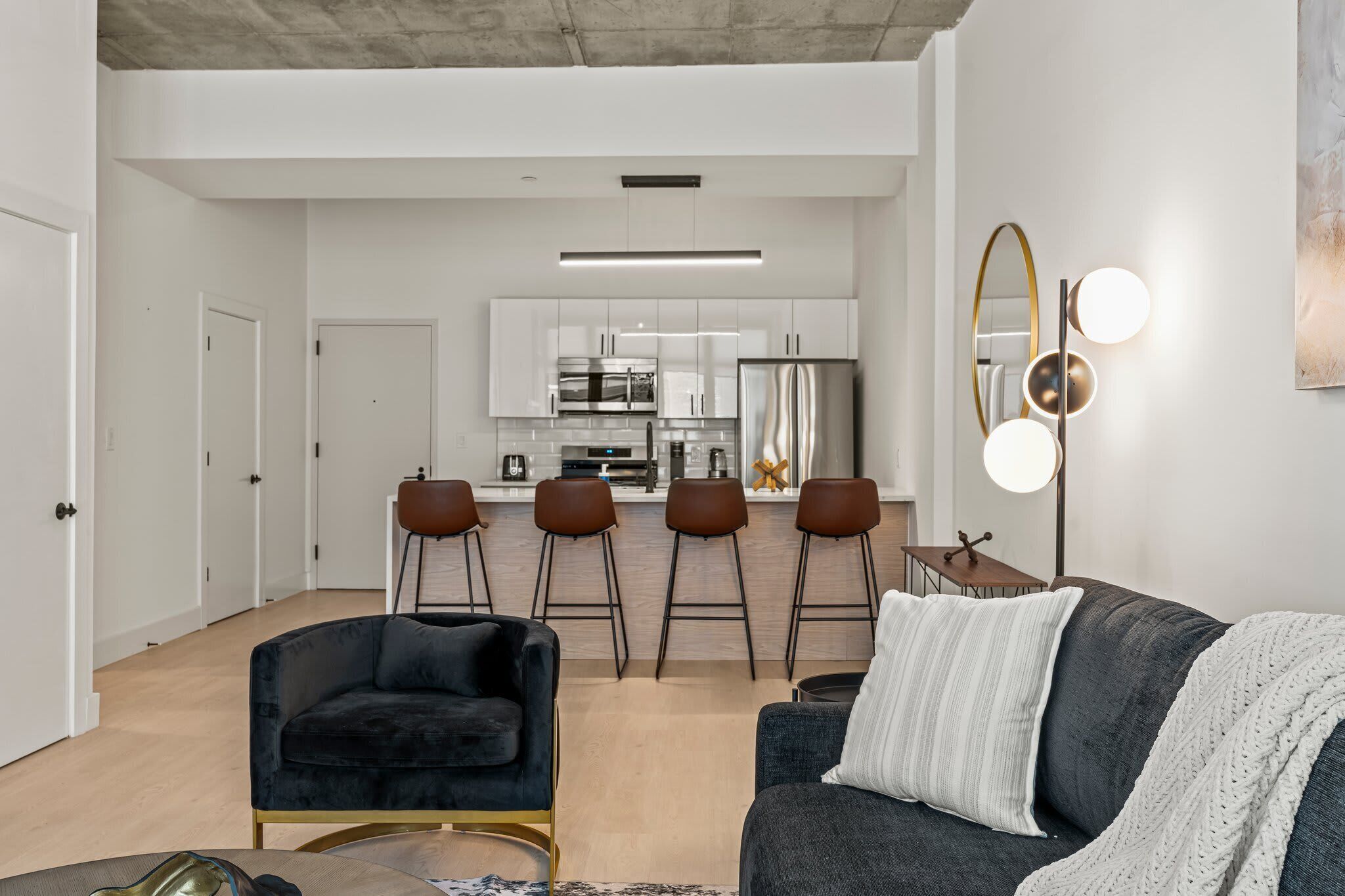 A modern living room with a dark sofa, a black armchair, and a coffee table. A kitchen with white cabinets and a bar area is visible in the background.