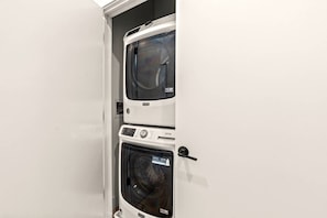 A white wall with a door partially open, revealing a white washing machine and dryer stacked vertically. - Sleek 1BR Sleeps 4 by NoLibs Waterfront Philly (Philadelphia)
