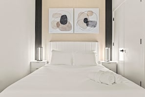 Ein modernes Hotelzimmer mit einem großen Bett, weißer Bettwäsche, zwei an der Wand befestigten abstrakten Gemälden und einem Nachttisch mit einer Lampe.