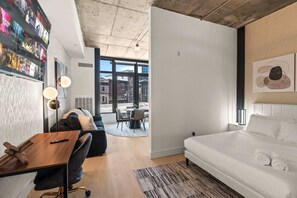 Ein modernes Hotelzimmer mit großem Fenster, einem Bett, einem Schreibtisch mit Stuhl, einer Couch und einem Essbereich mit Stühlen.
