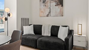 Ein modernes Wohnzimmer mit einem dunkelgrauen Sofa, weißen Kissen, einer Stehlampe und einem Beistelltisch mit Spiegel.