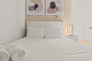 Un lit bien fait, avec du linge de lit blanc et des oreillers, une table de chevet avec une lampe, et une œuvre d’art abstraite accrochée au mur au-dessus du lit.