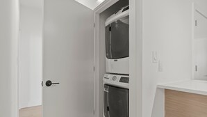 A laundry room with a stacked washer and dryer, a white door, and a wooden floor.