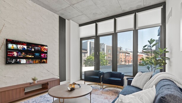 A modern living room with a large TV mounted on the wall displaying a Netflix interface, a round coffee table, a sofa, and a large window offering a view of the city.