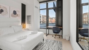 A modern hotel room with a large bed, a desk, and a view of the city through large windows.