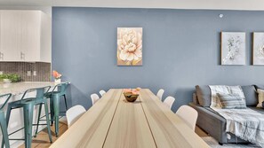 A dining area with a long wooden table, white chairs, a blue wall with floral artwork, and a grey sofa with a blanket. - Luxury 1BD | Uptown | Sleeps 4 | Parking (Dallas)