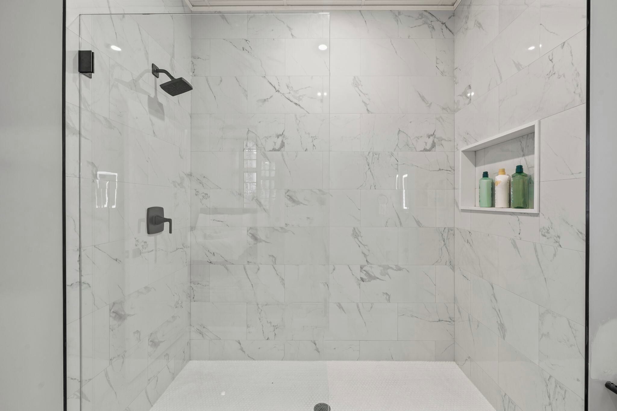 Un área de ducha moderna con puerta de vidrio, azulejos de mármol y un estante con artículos de aseo.