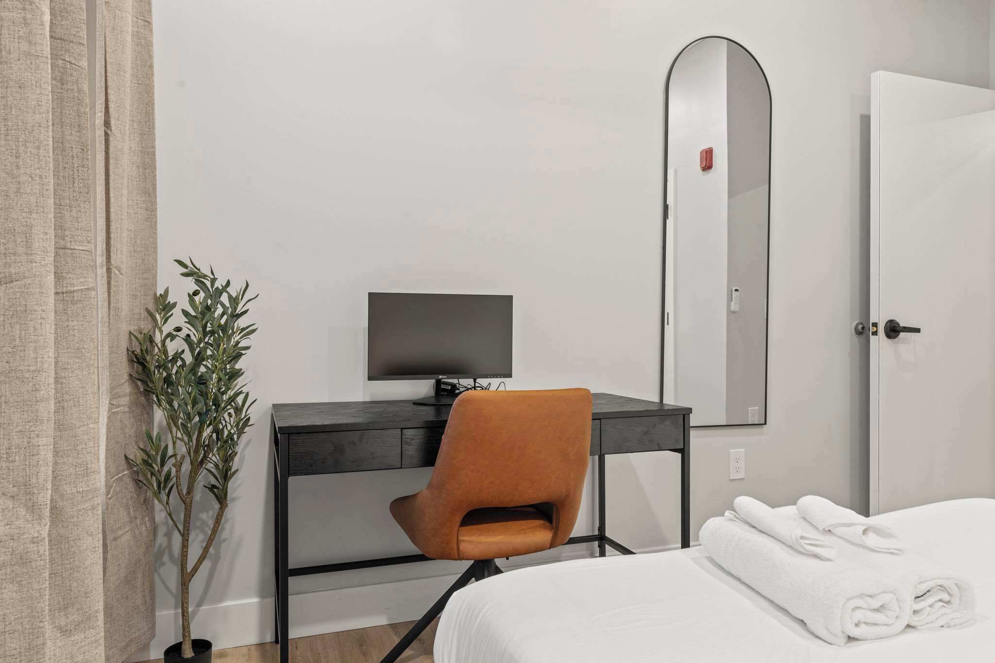 Una habitación de hotel con una cama, un escritorio con computadora, una silla, una planta en maceta y un espejo.