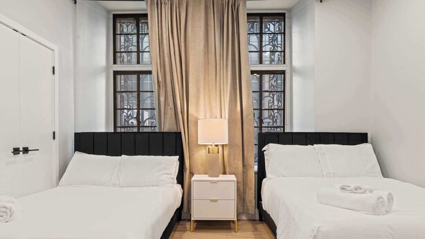Habitación de hotel con dos camas, una mesita de noche con lámpara, una ventana con cortinas y una puerta.