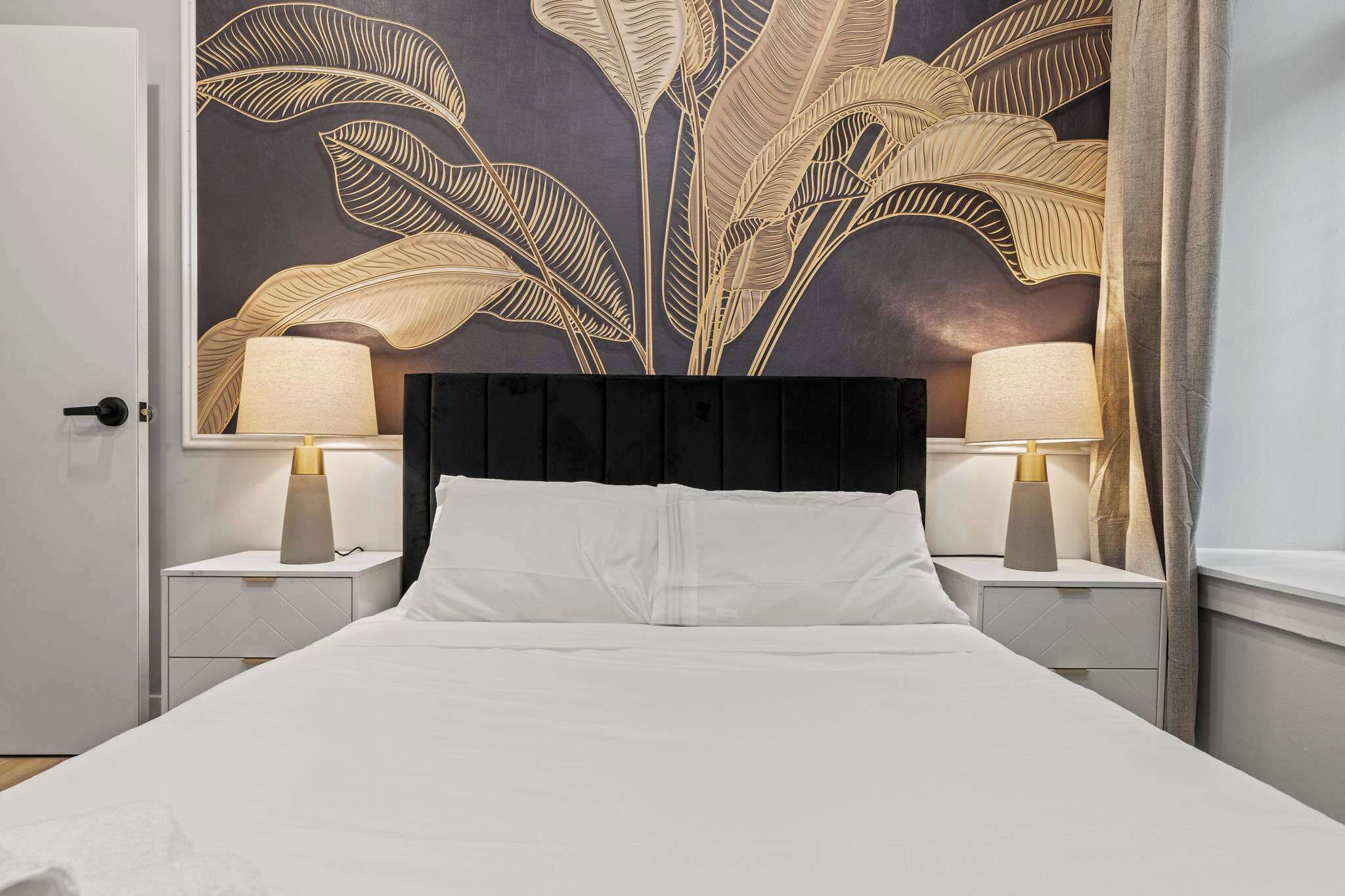 Habitació d'hotel amb un llit gran, dues taules de nit amb làmpades, un panell de capçal fosc i un mural de fulles a la paret.
