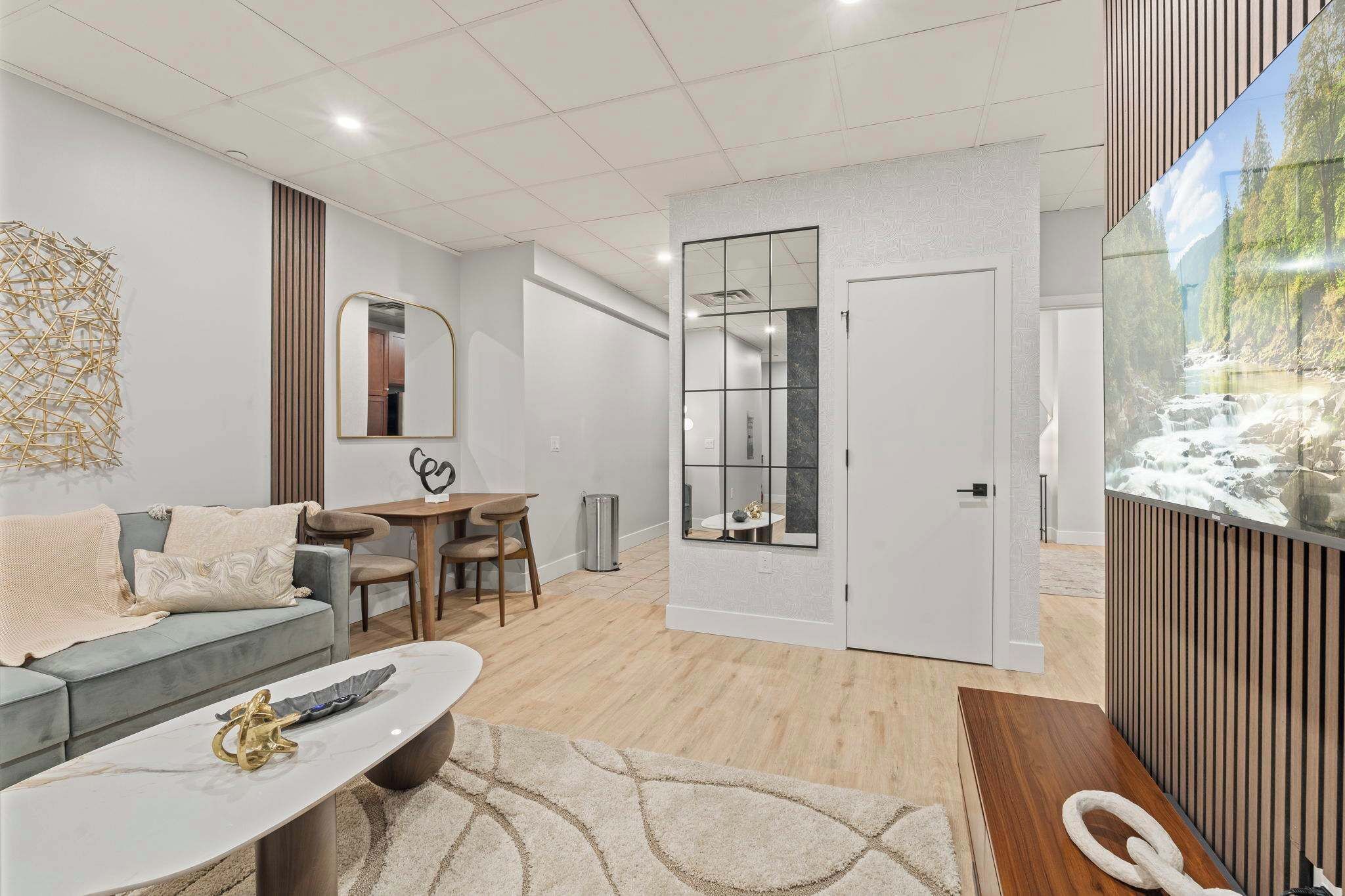 Un salon moderne avec un canapé gris, une table basse et un coin repas comprenant une table et des chaises. On y trouve également un grand miroir et une porte coulissante qui donne sur une autre pièce.
