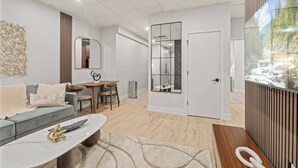 A modern living room with a grey sofa, a coffee table, and a dining area with a table and chairs. There is a large mirror and a sliding door leading to another room.