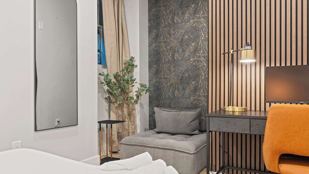 A modern hotel room with a bed, a grey sofa, a desk with a chair, a wall-mounted lamp, and a large mirror.