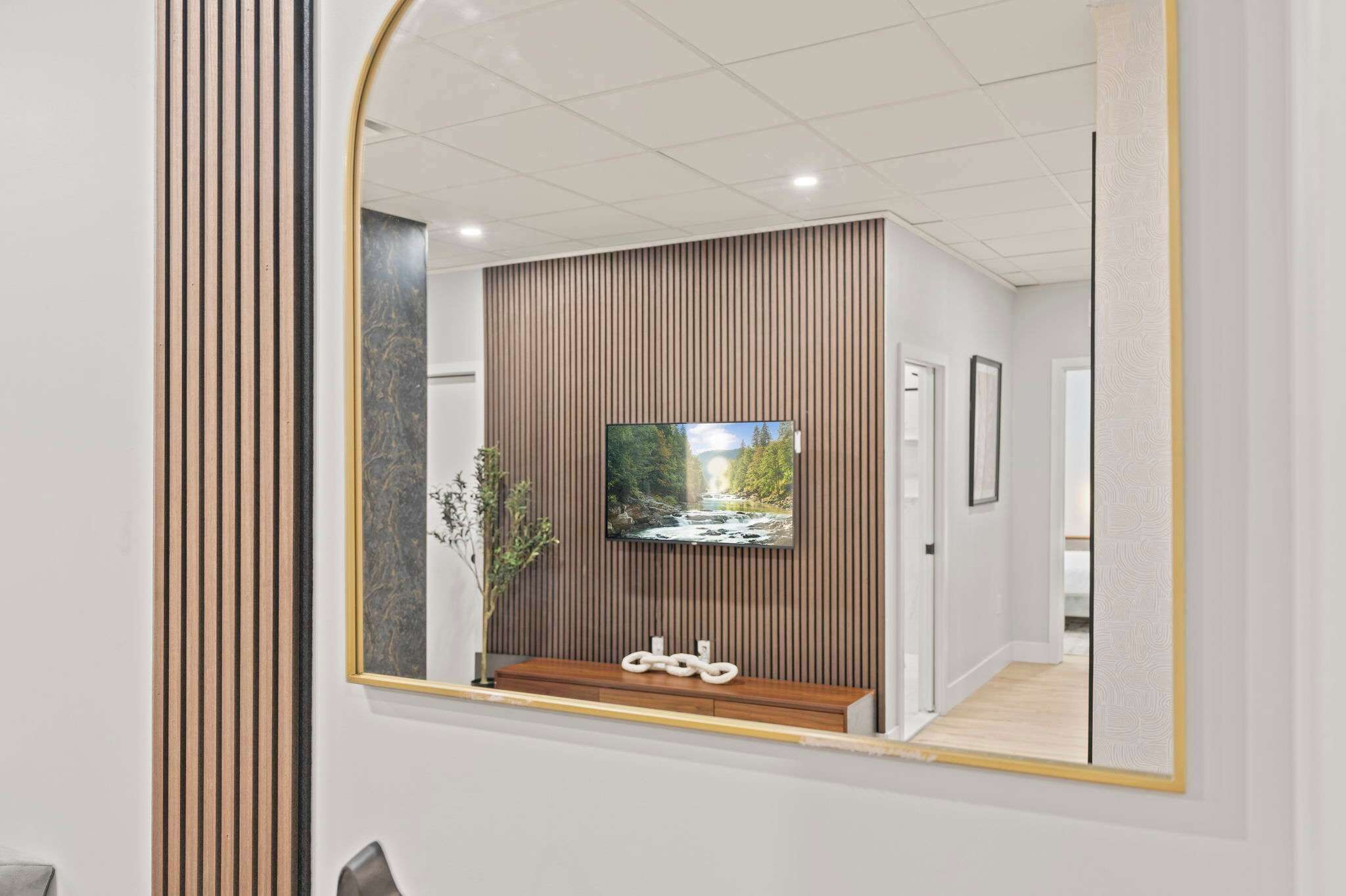 Un intérieur moderne avec un grand miroir qui reflète une pièce : on y voit un téléviseur fixé à un mur recouvert de panneaux en bois, un petit banc en bois et un tableau encadré.