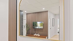 A modern interior with a large mirror reflecting a room with a TV mounted on a wooden panel wall, a small wooden bench, and a framed picture.