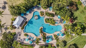 Vista aérea de un resort con una piscina grande y de forma irregular rodeada de palmeras, sillas de descanso y un pequeño edificio.