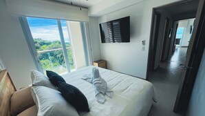 A hotel room with a large bed, a TV mounted on the wall, and a view of greenery through the window. - Spectacular View 3 bedroom Ocean / Rainforest View Condo With Beach Club Pass (Jaco)