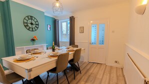 Un coin repas avec une table blanche dressée pour quatre personnes, une horloge au mur et une fenêtre avec des rideaux.