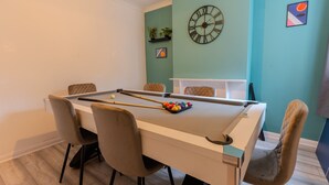 Une table de billard avec des chaises, une horloge et une étagère avec des plantes, le tout dans une pièce aux murs de couleur turquoise.