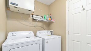 A laundry area with two white washing machines, a wall-mounted air conditioner, and a shelf with cleaning supplies. - Cozy, Comfortable and Spacious 2 Bedroom Home (Moncton)