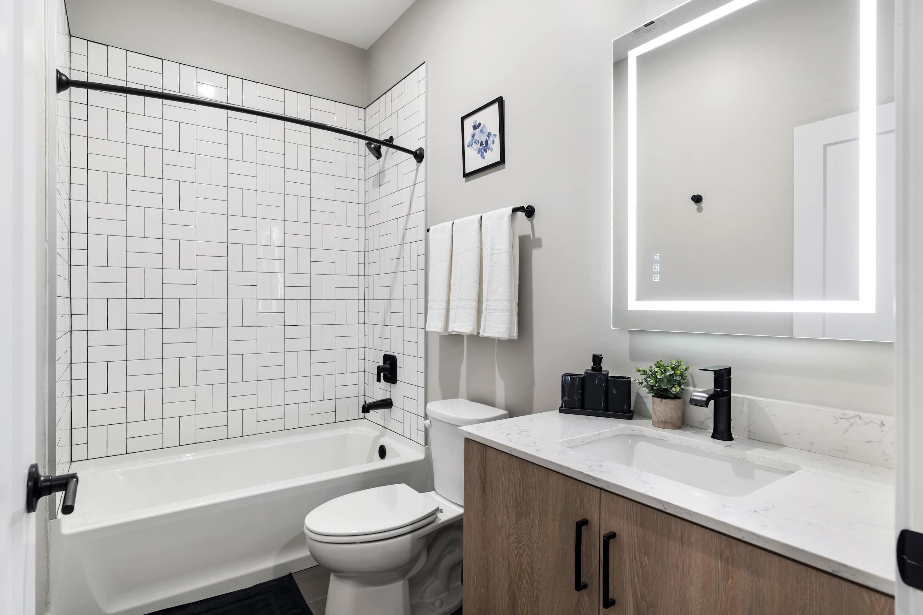 A modern bathroom with a white tiled shower, a white toilet, a white sink with a marble countertop, and a wooden cabinet.