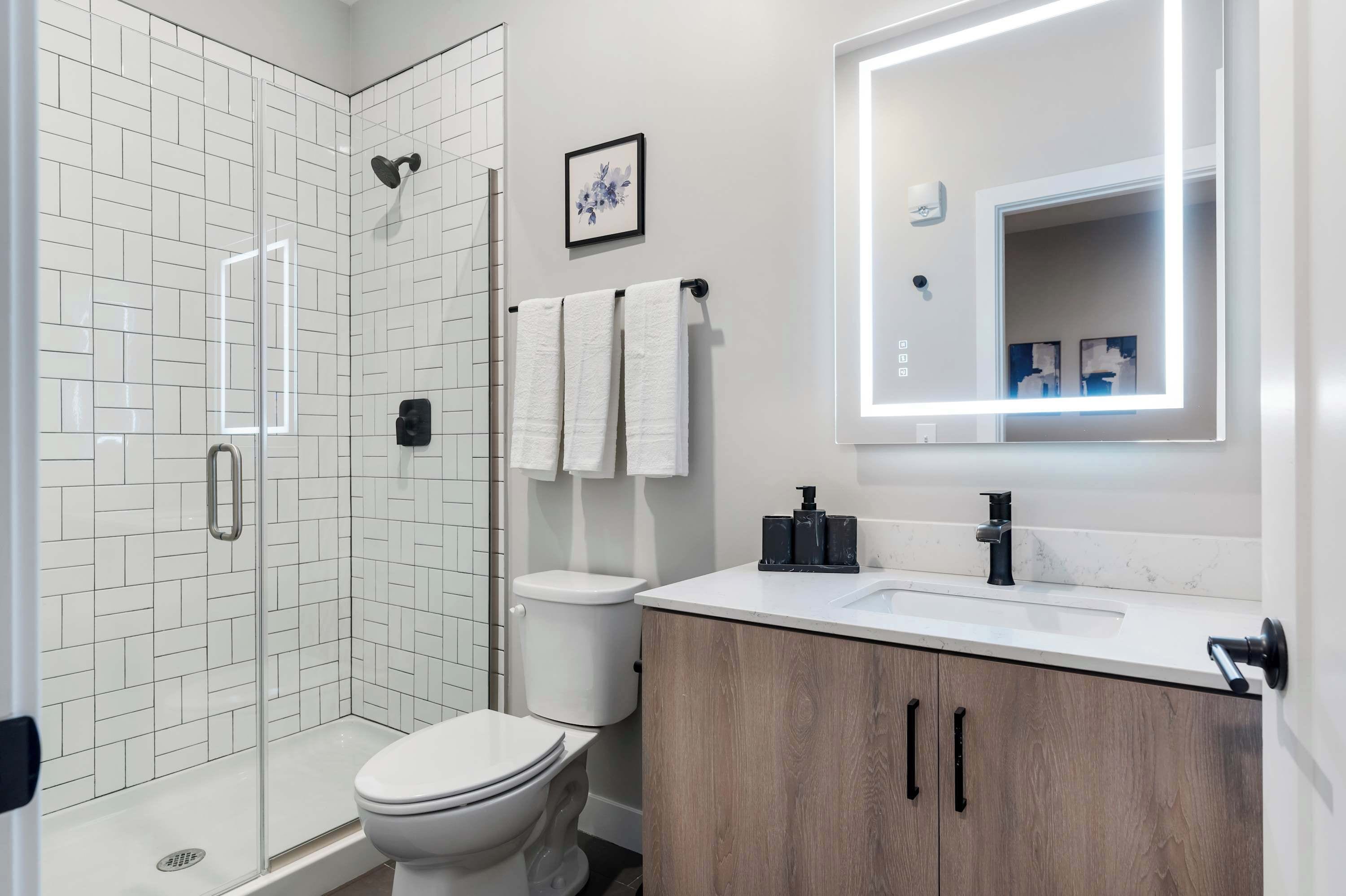A modern bathroom with a glass shower enclosure, white tiled walls, a white toilet, a wooden vanity with a black faucet, and a large mirror with built-in lighting.