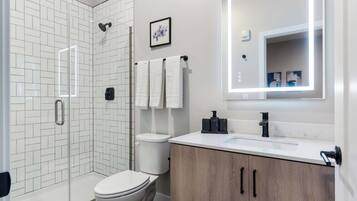 A modern bathroom with a glass shower enclosure, white tiled walls, a white toilet, a wooden vanity with a black faucet, and a large mirror with built-in lighting.