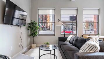 Una sala moderna con un sofá, una mesa redonda de centro, un televisor montado en la pared y ventanas amplias con vistas a la ciudad.