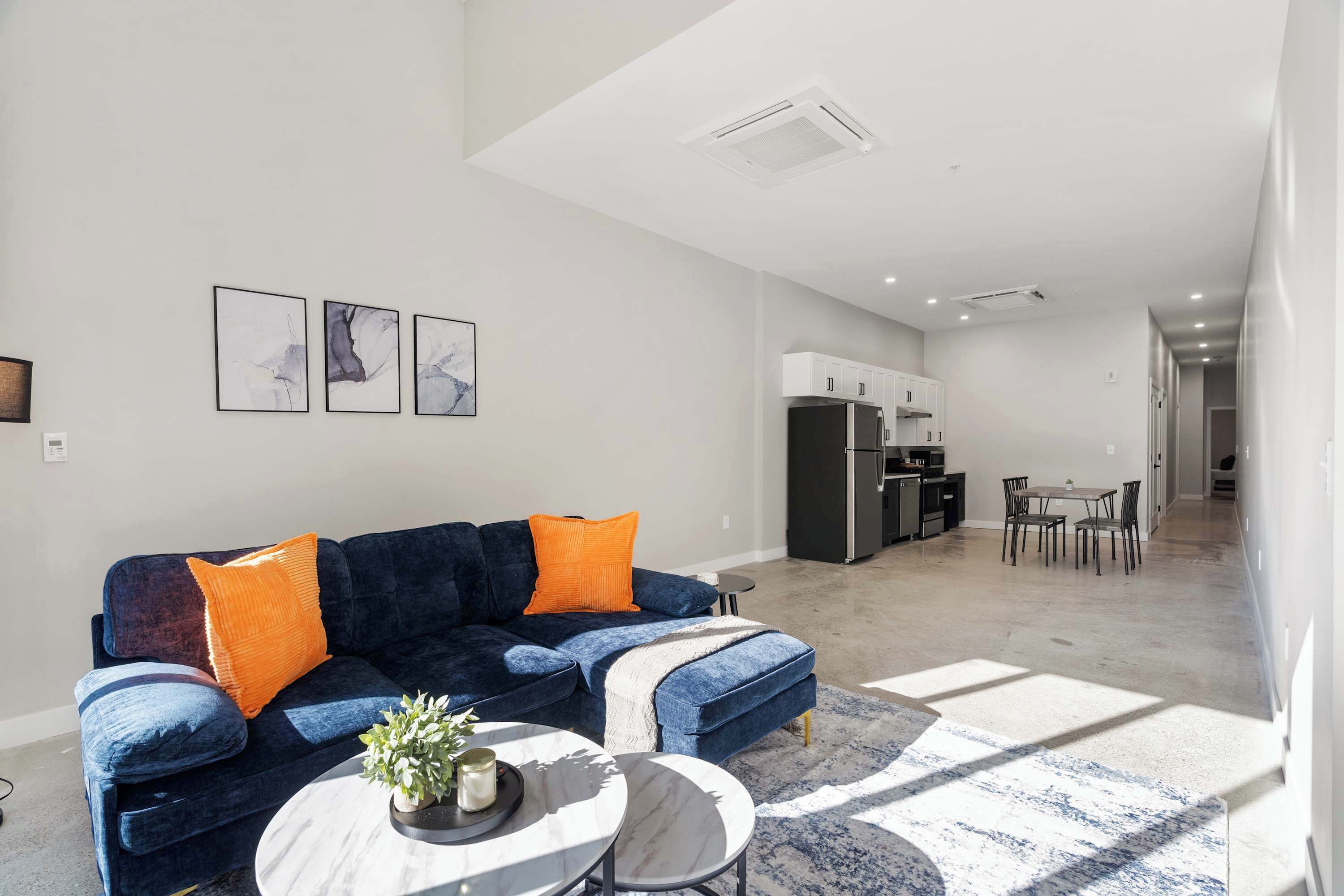 A modern living room with a dark blue sofa, two orange cushions, a marble coffee table, and a dining area in the background.