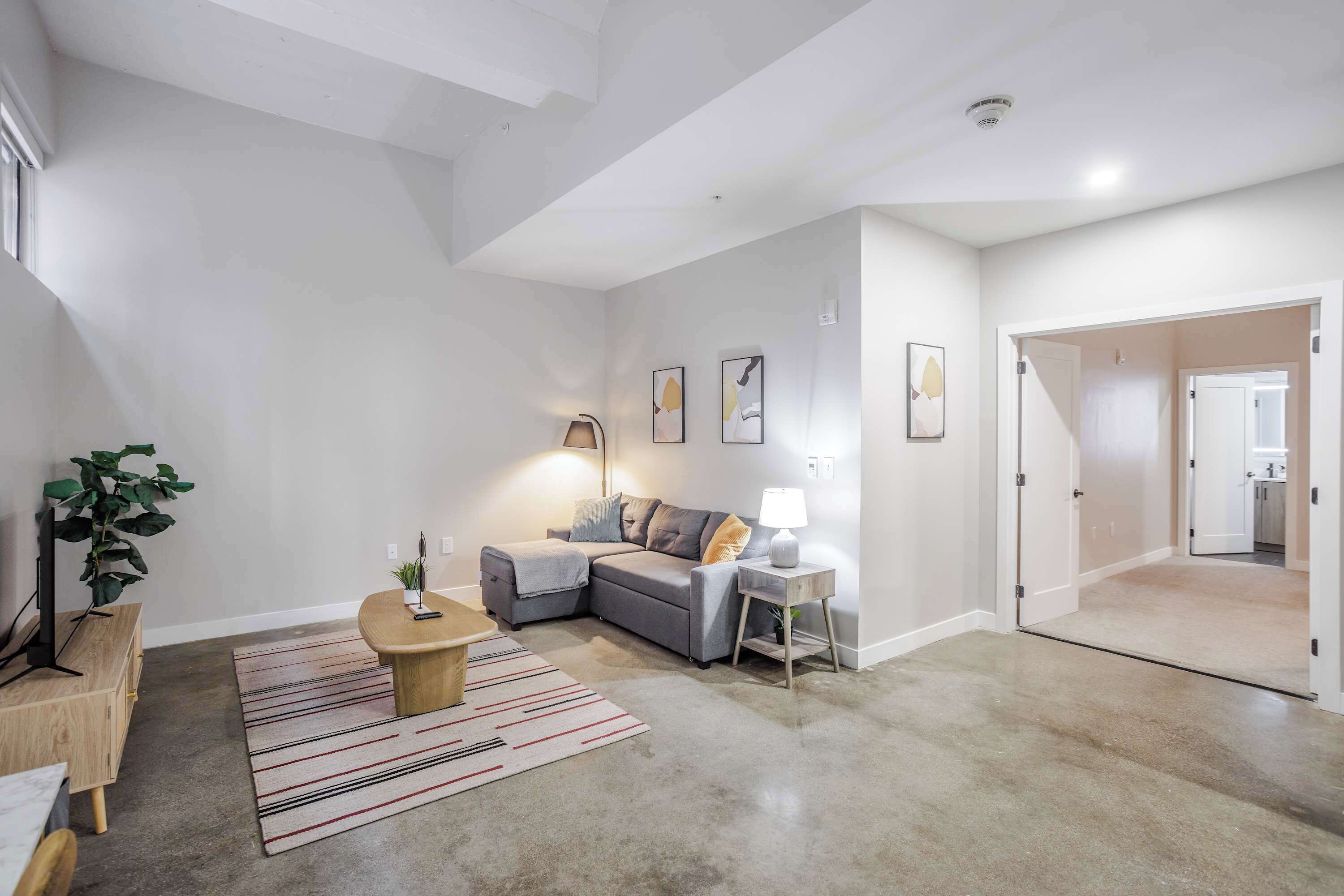 A modern living room with a grey sectional sofa, a wooden coffee table, and a striped rug. There is a lamp on a side table, and a plant in the corner.