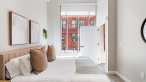 A bedroom with a bed, pillows, a bedside table, and a window with a view of a building. - CozySuites at Textile (Cincinnati)