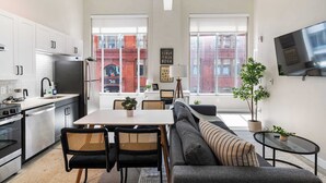 A modern kitchen with white cabinets, a stainless steel refrigerator, and a dining area with a table and chairs. There is a large window providing a view of the city. - CozySuites at Textile (Cincinnati)