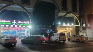 Una entrada de hotel con un gran arco, varios coches aparcados delante y un cartel que indica AMAL AL MASHAAER HOTEL.