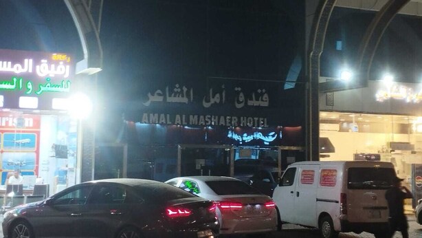 Entrada de hotel con señalización en árabe e inglés, varios vehículos estacionados frente y una acera para peatones.