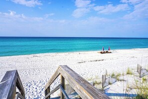 Ein Strand mit einer Holzpromenade, die zum Wasser führt, einem sandigen Ufer und einem klaren, blauen Himmel.