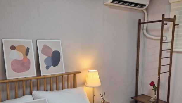 Una habitación de hotel ordenada con cama, una mesita de noche con lámpara, un estante pequeño con un florero y un equipo de aire acondicionado en la pared.