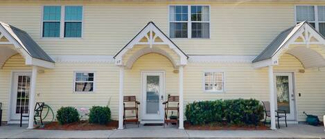A row of yellow buildings with white trim, numbered 17 and 18, featuring a central door with a small porch and two wooden chairs.