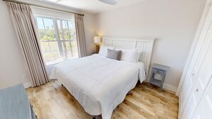A bedroom with a large bed, a bedside table with a lamp, a window with curtains, and a view of trees outside.