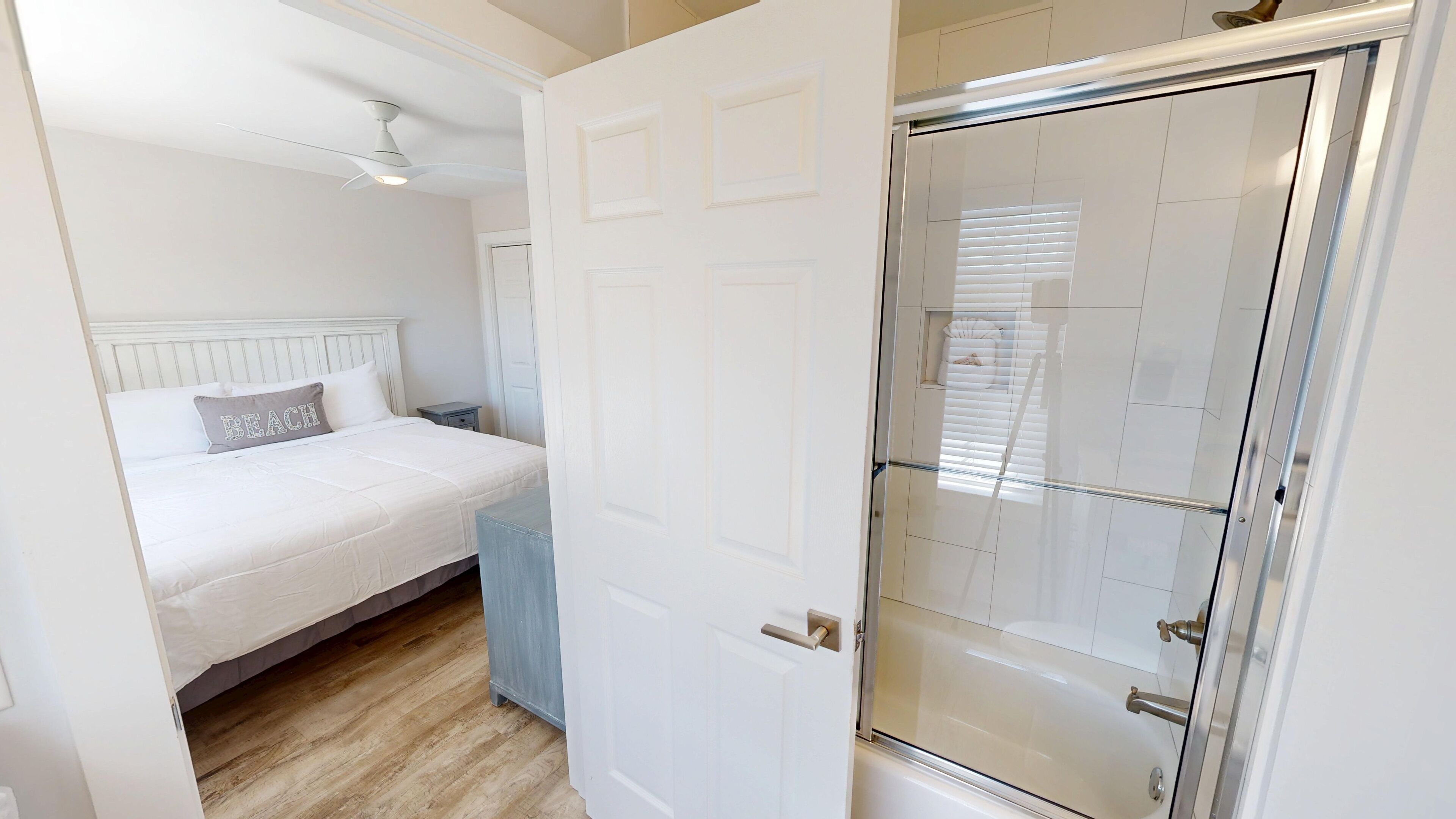 A bedroom with a bed, a nightstand, a ceiling fan, and a bathroom with a glass shower door.